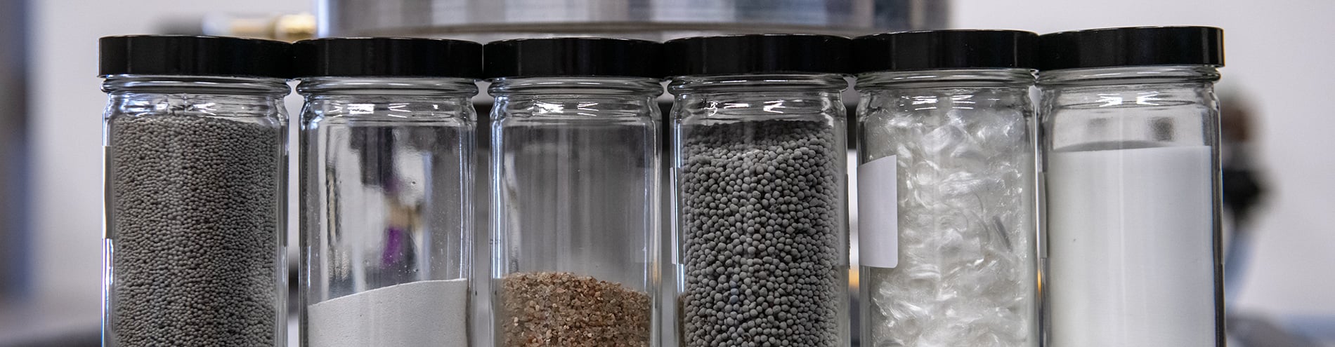 Left to right line of six clear glass jars with black lids - each holding a different dry industrial material.