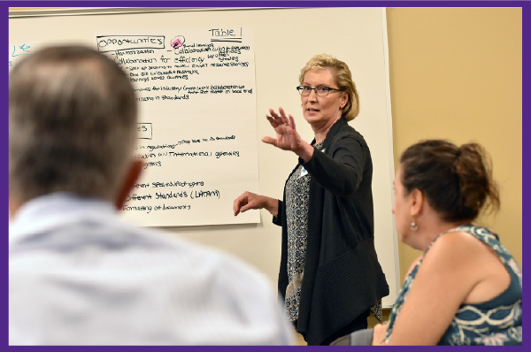Woman with short blonde hair gestures as she leads a classroom training session.